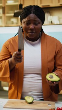African American woman records cutting avocado in food vlog at kitchen counter, influencer filming healthy breakfast idea indoors. Female influencer explaining meal prep and recipe development.