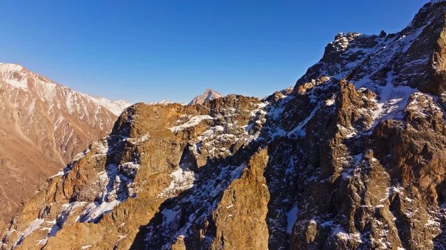 Majestic brownish sedimentary snow-capped mountains rise against a clear blue sky in Kyrgyzstan. Dolly forward drone view.