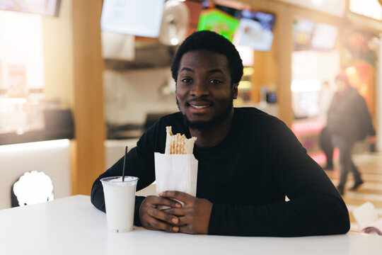 Young african male enjoying snack shaverma with chicken and drink at modern food court