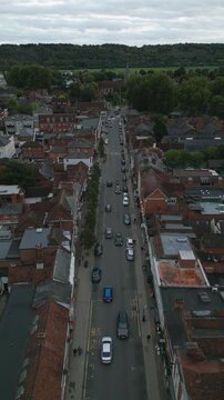 Aerial view of a town street lined with brick buildings, cars, and trees, with a lush forest in the background.