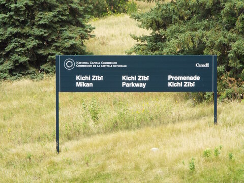 Ottawa, Ontario, Canada-July 23, 2025: A "Kichi Zībī Mīkan" parkway sign by the National Capital Commission stands in a grassy field bordered by evergreen trees.