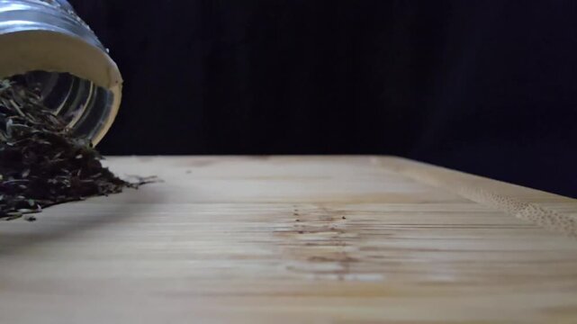 Close-up of dried thyme being poured from a glass jar onto a wooden cutting board. The movement captures the falling herbs and earthy textures under warm studio light against a black background.