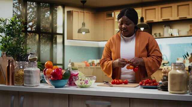 African American food blogger filming meal prep tutorial with cherry tomatoes in her home kitchen. Solo content creator preparing vegetables and other ingredients for a salad dish.