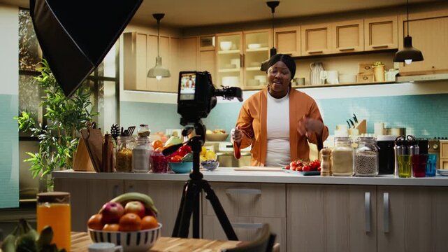 Recording on tripod, African american woman, vlogging culinary content, filming behind the scenes in home studio kitchen. Woman blogger ensuring meal preparation for online audience.