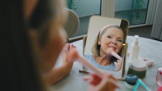 Elderly woman applying makeup in a bright room in the afternoon with a small mirror and beauty products on the table in front of her