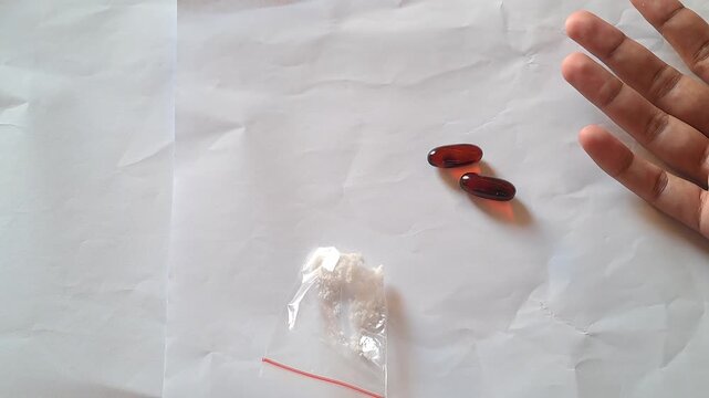 Close-up of Asian male hands demonstrating the dangers of illegal drugs using capsules and a small plastic bag of white powder as visual aids for drug abuse prevention education.