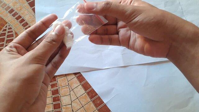 Close-up of Asian male hands demonstrating the dangers of illegal drugs using capsules and a small plastic bag of white powder as visual aids for drug abuse prevention education.