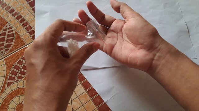 Close-up of Asian male hands demonstrating the dangers of illegal drugs using capsules and a small plastic bag of white powder as visual aids for drug abuse prevention education.