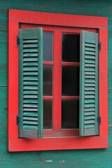 Obraz premium red-framed window with partially open weathered green shutters on a green wooden wall, rustic charming cottage detail