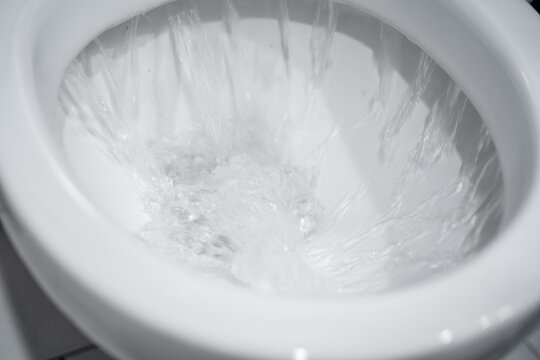 Top view into a toilet bowl as water flushes and splashes, white ceramic rim framing swirling clear water, bathroom sanitation concept with motion blur, reflections, clean minimal tones, turbulence
