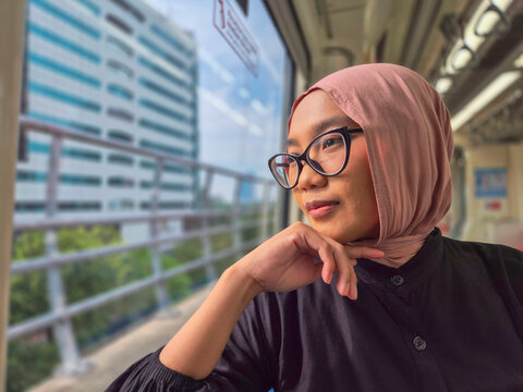Young Asian Muslim woman wearing a pink hijab and glasses, looking thoughtfully out the window while commuting on a modern city train