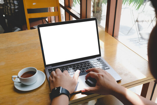Over the shoulder view of a person typing on a laptop with a blank white screen at a wooden table in a cafe next to a cup of coffee.