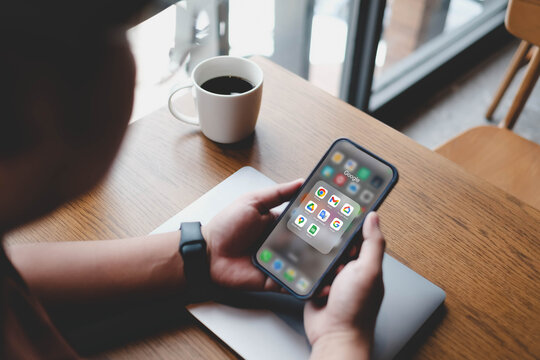 Bangkok, Thailand. MAR 23, 2026; A person holding a smartphone displaying a folder of Google applications, including Chrome, Gmail, and Drive, while sitting at a wooden table with a coffee cup.