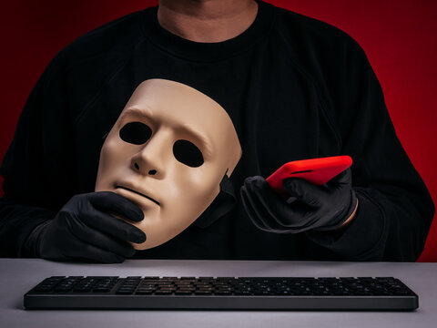 Online scam concept showing anonymous criminal or hacker wearing gloves with mask and smartphone impersonating. Phishing attack, identity theft, digital fraud and cybercrime threat on red background.