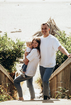 The photograph depicts a family standing on a staircase leading to the seashore. The man and woman are hugging each other, and a small child is nearby. Everyone looks happy and relaxed, enjoying their