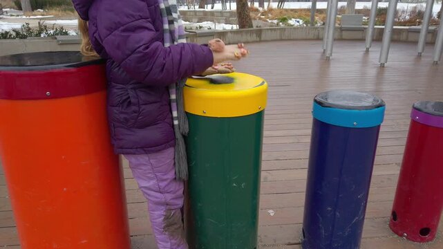 A young girl in a purple winter jacket and scarf plays on vibrant, colorful percussion tubes in an outdoor musical playground, developing rhythm and motor skills in a public park.