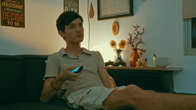 Caucasian man sitting on a sofa in his living room, looking bored while channel surfing with a remote control during the evening