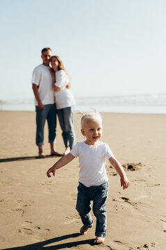 A small child walks barefoot along a sandy beach. He smiles and looks very happy. In the background, his parents stand, hugging each other and watching his child. The photograph conveys a sense of fam