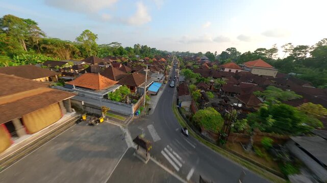 Cinematic FPV drone flight over traditional village in Bali, Indonesia, starting above local temple and continuing along decorated street surrounded by rice fields, lush greenery and village houses.