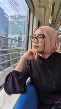 Young Asian Muslim woman wearing a pink hijab and glasses, looking thoughtfully out the window while commuting on a modern city train