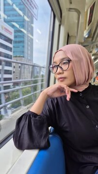 Young Asian Muslim woman wearing a pink hijab and glasses, looking thoughtfully out the window while commuting on a modern city train