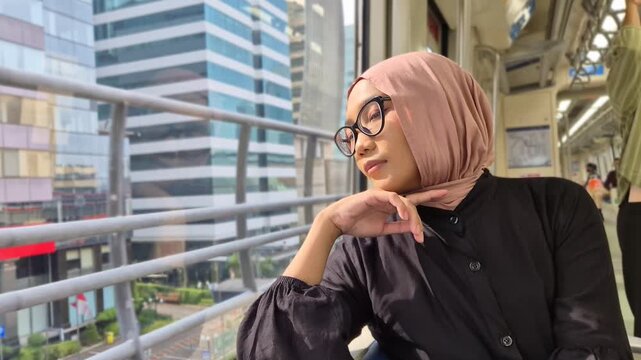 Young Asian Muslim woman wearing a pink hijab and glasses, looking thoughtfully out the window while commuting on a modern city train
