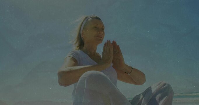 Senior woman breathing, bringing hands into prayer-pose on sand with headband bracelet for wellness