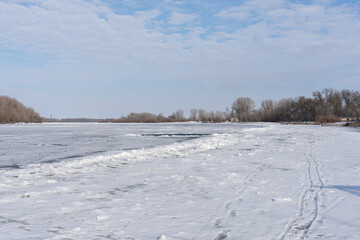 Obraz premium Expansive winter landscape of a frozen river covered in snow and ice. Features an ice-hole, tracks in the foreground, and a distant shore with bare trees under a pale blue cloudy sky.