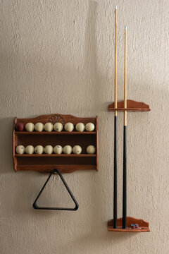 A still life of classic billiard equipment. Ivory balls and two cues are stored in wooden wall racks against a beige textured background. A black triangle hangs ready for the next game.