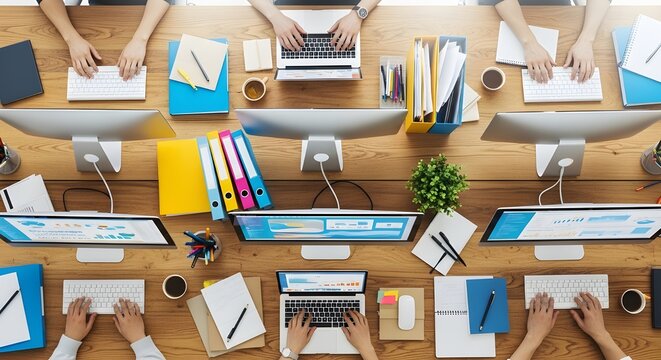 Overhead view of collaborative office workspace with people working on laptops and computers surrounded by office supplies and paperwork on a wooden desk