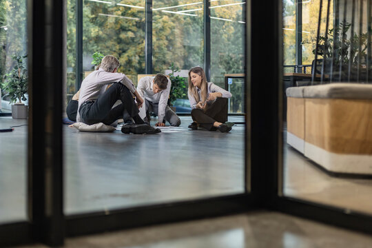 Business people looking at project plan laid out on floor, discussing new project plan in modern office, view from glass wall