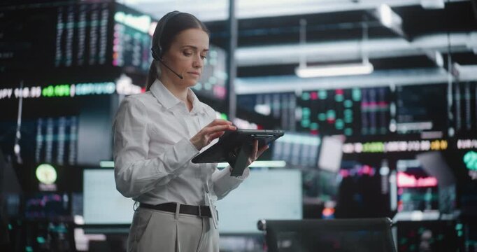 Professional Female Stock Exchange Trader on Trading Floor With Tablet, Monitoring Live Market Data on Digital Screens. Wearing Headset, Analyzes Stock Fluctuations and Coordinates Trading Operations.