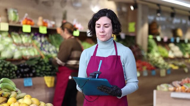 Woman store employee with documents takes inventory of goods on display case of vegetable store. Seller records quantity of goods in report, draws up document on availability of vegetables in store