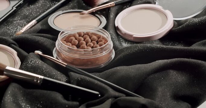Rotating professional makeup set with various products including bronzing pearls, highlighter, face powder, and brushes lying on a luxurious black sparkling fabric for a glamorous look