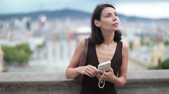 Thoughtful woman in an elegant dress stands in front of a European city, she walks in the summer with a mobile phone and a small handbag in hands. Woman walking around Barcelona