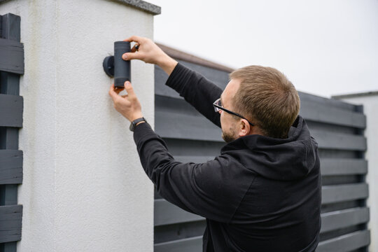 Adult man in casual clothes mounts outdoor wall lamp using screwdriver. Home improvement work, maintenance and installation in backyard of modern house.