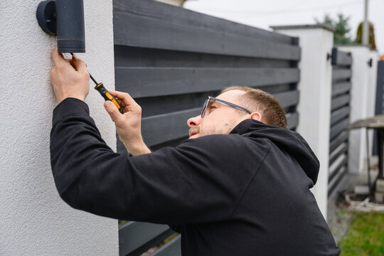Adult man in casual clothes mounts outdoor wall lamp using screwdriver. Home improvement work, maintenance and installation in backyard of modern house.