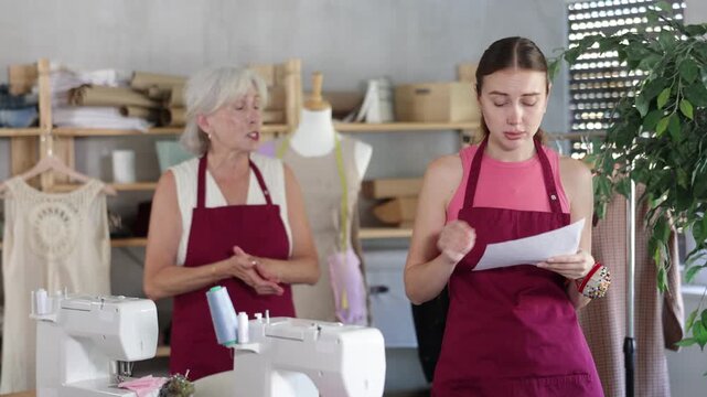 Elderly female tailor scolding adult female assistant with document for dismissal in sewing workshop