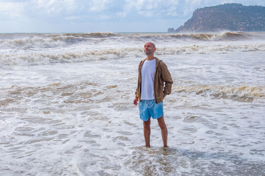 happy positive caucasian Bald mature man walks barefoot in foamy surf, rough waves background, stormy weather, storm warning, enjoys solitary walk powerful sea, beach closed swimming, mental health