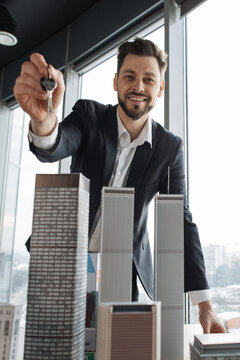 A smiling agent presents keys over a scale model of high-rise buildings in a modern office setting with city views and daylight windows indoors