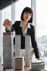 A smiling woman in a suit holds keys in front of miniature skyscraper models, symbolizing property...