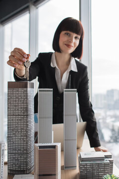 A smiling woman in a suit holds keys in front of miniature skyscraper models, symbolizing property ownership and development
