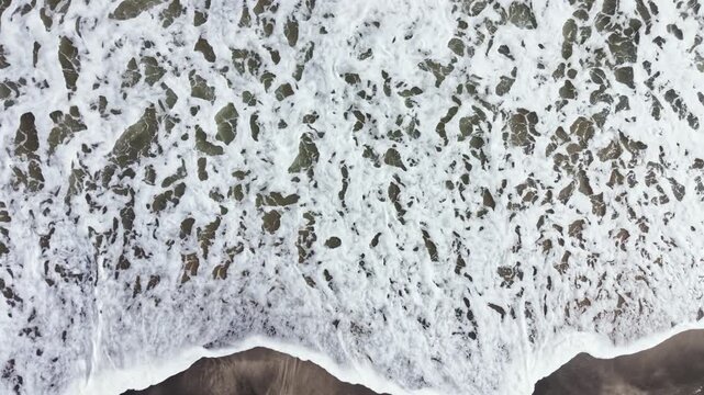 Waves crash against the shore on a sandy beach under bright sunlight. The foam from the water creates patterns as it meets the sand. This scene shows nature's power and beauty.