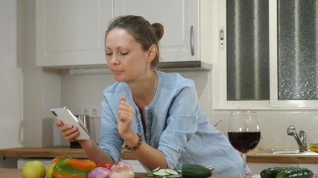 Positive casual lady relaxing with red wine while checking social media on the phone at the kitchen