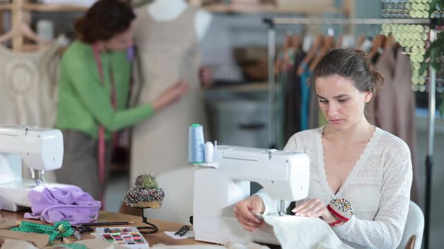 Woman designer sews clothes using sewing machine, perform edge finishing stitching seam. Companion work in background with mannequin
