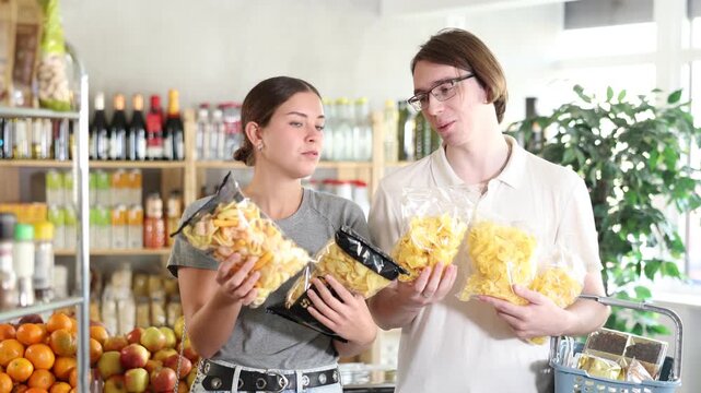 Youth guy with wife choose and buy chips and potato mix crisps in store. Visitors make choice of assortment, buy potato snack crispy rings, sticks. Clients hesitate before buying