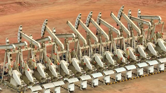 Drone perspective capturing multiple oil pump jacks operating in a barren landscape, symbolizing energy demand, petroleum extraction, and the mechanics behind fuel production systems.
