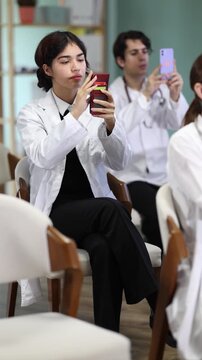 Positive young malaysian female medical student sitting and listening lecture, taking photo or QR with mobil or tablet in light lecture hall