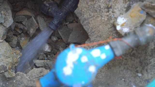 Water gushes from a ruptured underground pipe in a rocky excavation pit. Professional plumbing repair site with a blurred blue water meter in focus. Infrastructure failure and utility emergency.