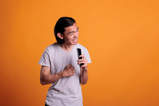 Young asian man singing karaoke, grimacing, using smartphone as microphone. Singer practicing song, teenager holding mobile phone as mic, creative hobby, fun pastime leisure activity